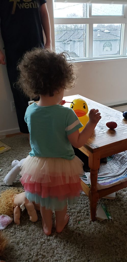 A curly-haired toddler wearing a tulle skirt paints the eyebrow of a very large rubber duckie.