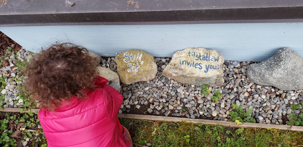A toddler in a pink coat bends down to investigate two rocks, one of which is painted with "Still, sit." and the other "tastatlaw invites you."