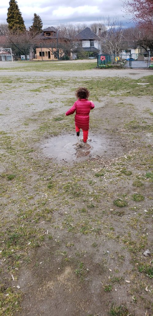A toddler in a pink coat runs in a mud puddle in only her sock feet.