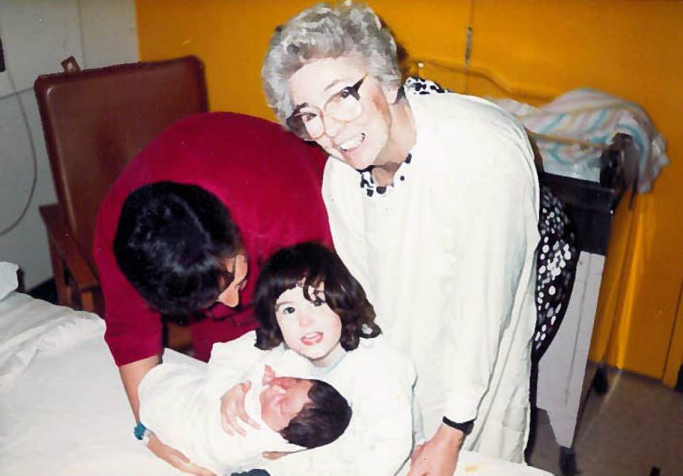An elderly woman with big glasses, a woman wearing maroon bends over to show a dark-hair two-year-old a new baby