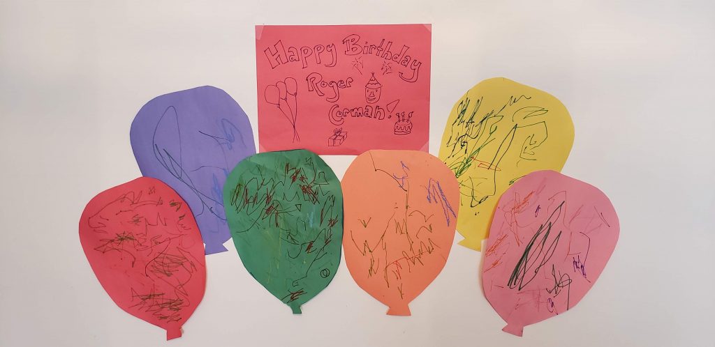 Red sign that reads "Happy birthday roger corman." 6 construction paper balloons decorated with toddler scribbles