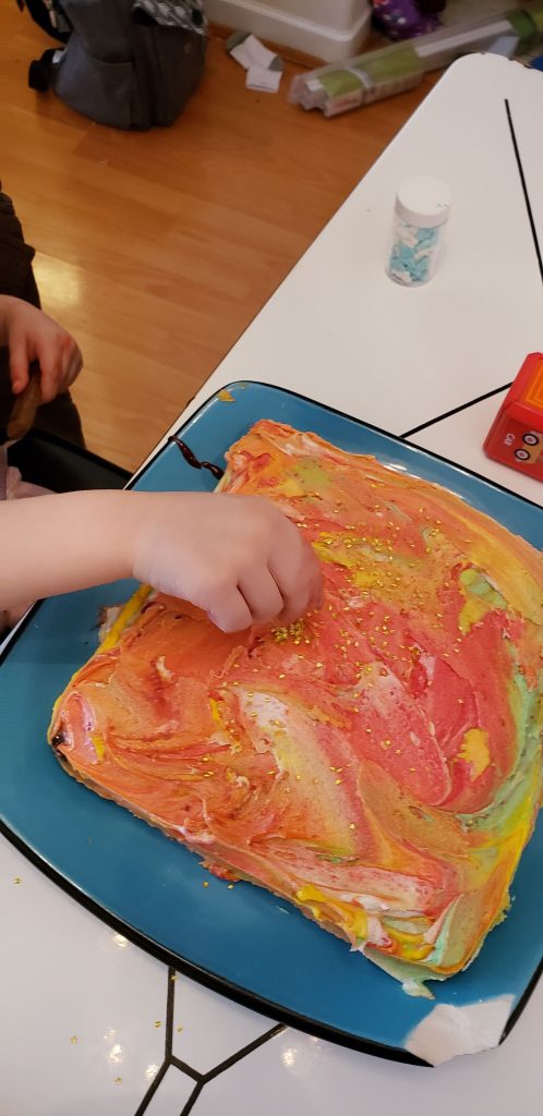Toddler puts gold sprinkles on a rainbow cake on a blue plate