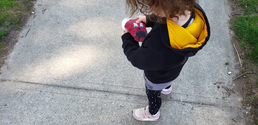 A toddler in a black and yellow hoodie holds a rock that's been painted maroon with a face on it.